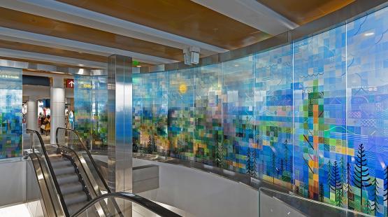 Glass panels curved around an escalator, painted with a pixelated landscape of trees and mountains. 