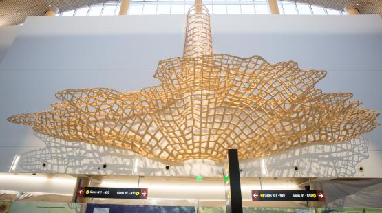 A large cantilevered wood sculpture spanning the ceiling of SEA Airport North Satellite concourse, viewed from below.