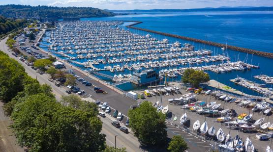Aerial view of a large marina filled with rows of docked boats and yachts beside a long breakwater extending into a blue body of water. A parking area, trees, and additional sailboats on trailers are visible in the foreground, with forested hills and a partly cloudy sky in the background.