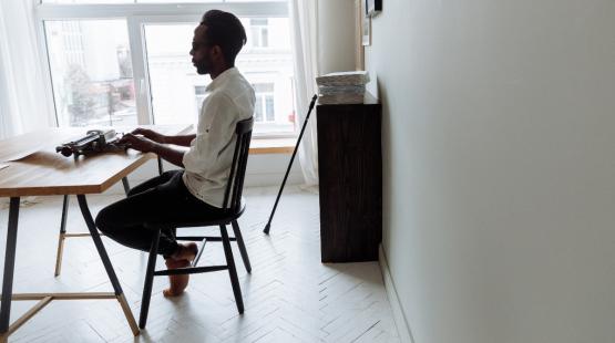 Person seated at a desk typing on a typewriter in a bright room, with a window behind the desk and a walking cane leaning against a cabinet.