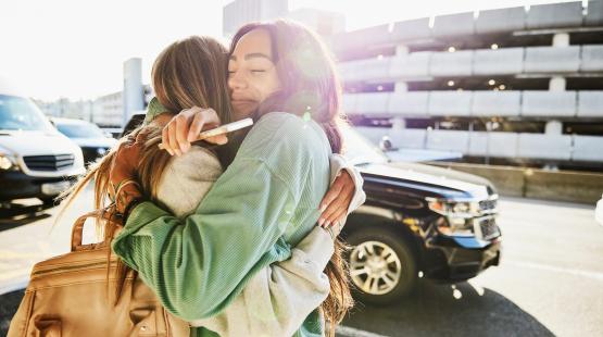 Two people embracing on an airport curbside area, with vehicles and a multi‑level parking structure in the background. One person is holding a mobile phone while carrying a backpack.