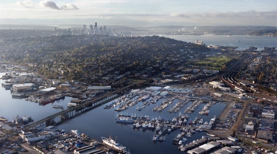 Aerial view of a large waterfront area with numerous docks and boats in the foreground, a densely built residential neighborhood on a hillside in the middle ground, and a downtown skyline with tall buildings and the Space Needle in the background under partly cloudy skies.