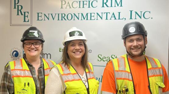 Three people wearing safety vests and hard hats standing in front of a sign for “Pacific Rim Environmental, Inc.