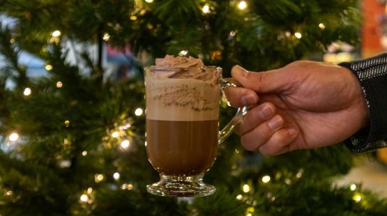 Hand holding a glass mug of a whipped‑topped coffee drink in front of a lit holiday tree.
