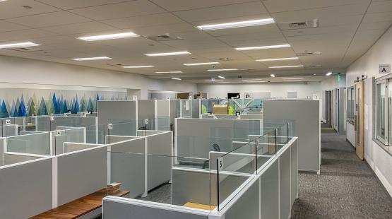 A large indoor office space with rows of light gray cubicles separated by glass panels, each labeled with numbers. The area is well-lit with ceiling lights, and a hallway runs along the right side of the room. A wall on the left features a decorative mural with blue and green triangular shapes.