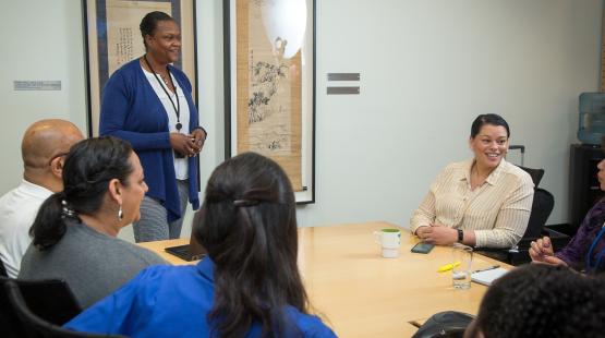 A group of people seated around a conference table while another person stands at the front of the room speaking. The room has framed artwork on the wall and a water dispenser in the corner.