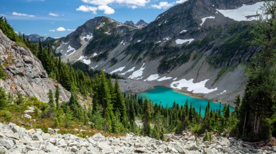 Mountain landscape with a turquoise alpine lake surrounded by rocky slopes, evergreen trees, and patches of snow under a blue sky.