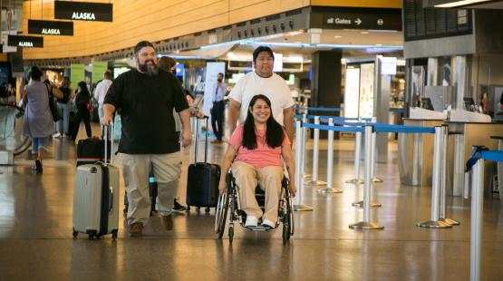 “Interior airport scene showing a person using a wheelchair being assisted by two people, all moving through a terminal area with luggage. Overhead signs display airline information, and stanchions form queue lines in the background.