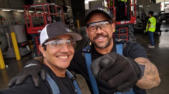 Two workers wearing safety gear take a selfie inside a warehouse, with lift equipment and additional workers visible in the background.