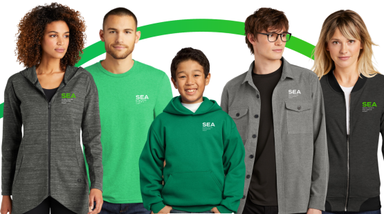 Five individuals stand in a row wearing SEA‑branded apparel, including zip‑up jackets, a bright green long‑sleeve shirt, a green hoodie, a gray button‑up shirt, and a dark gray jacket. A curved green graphic runs across the background behind them. The SEA logo is visible on each clothing item.