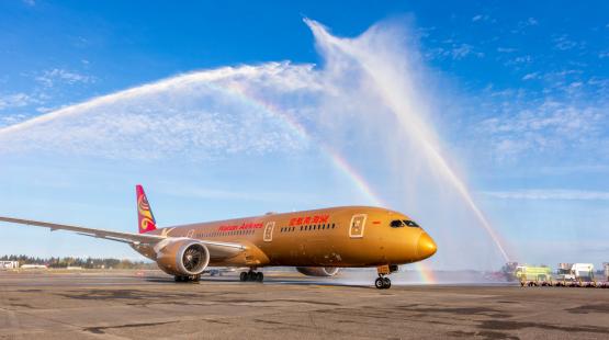 A gold-colored commercial airplane on an airport runway passes beneath crossing arcs of water sprayed by fire trucks, creating a visible rainbow against a bright blue sky.