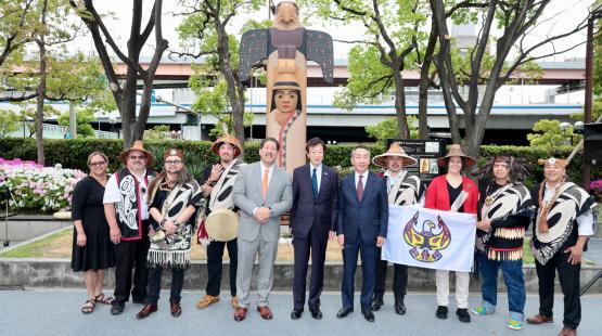 Group of people standing outdoors in front of a large carved wooden totem-like figure, surrounded by trees and landscaping. Several individuals wear traditional woven garments and hats, while others wear formal suits. One person holds a drum, and another holds a flag featuring a stylized bird design. A roadway and buildings are visible in the background.