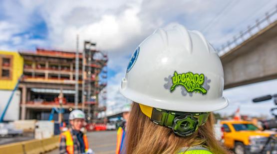 Worker with a construction hat that has the Upgrade SEA logo while a construction project is in progress in the background. 