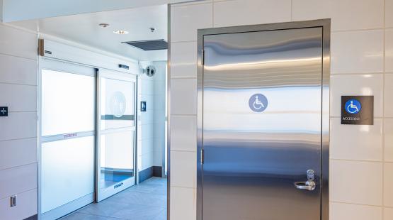 An accessible restroom entrance with a metal door displaying an accessibility symbol, next to frosted sliding doors leading to a restroom area.