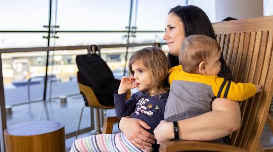 Person seated in a wooden chair at an airport gate area holding two young children on their lap.