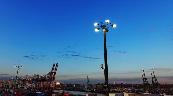 A tall pole with bright lights stands over a shipping port with cranes and containers at dusk.