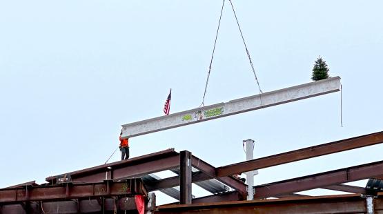 A steel beam with an American flag and a small evergreen tree is lifted by a crane onto a building frame, while a worker stands on the structure guiding it.