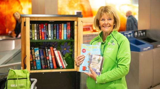 Becky Nickels stands beside a small wooden book-sharing cabinet and holds a few books and magazines.