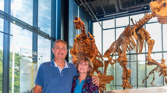 Two people stand in front of a large prehistoric animal skeleton displayed in a museum with tall windows behind them.