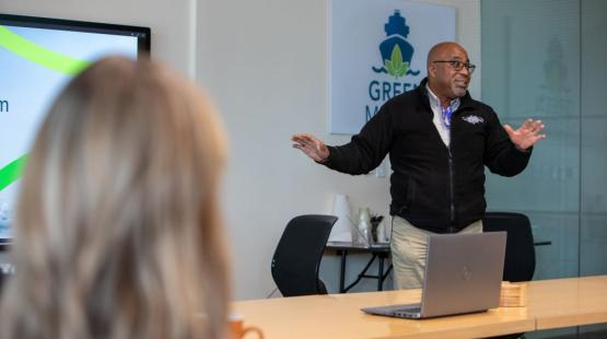 Delmas Whittaker stands at the front of a meeting room giving a presentation, with a laptop on the table and a screen displaying slides in the background.