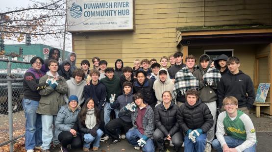 A large group of people pose together outside a building with a sign that reads “Duwamish River Community Hub.” Trees, a fence, and a street intersection are visible in the background.