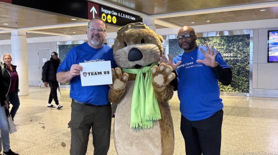 Two Boeing Cares volunteers stand in an airport with JETT, an otter mascot wearing goggles and a green scarf. One volunteer holds a “TEAM US” sign.
