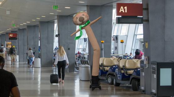 An airport concourse with gate signs and travelers, featuring a tall inflatable otter‑themed tube figure near seating and carts.