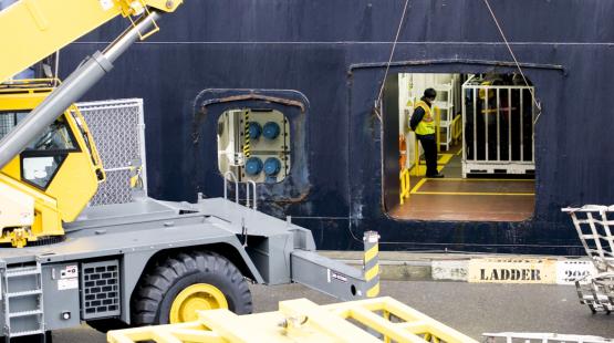 Cargo operation scene showing a large yellow crane positioned beside an open doorway on a ship, with a worker in a safety vest standing inside the vessel.