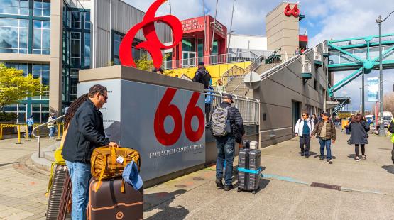 People with rolling luggage walk near a waterfront terminal building marked with a large red “66” sign.