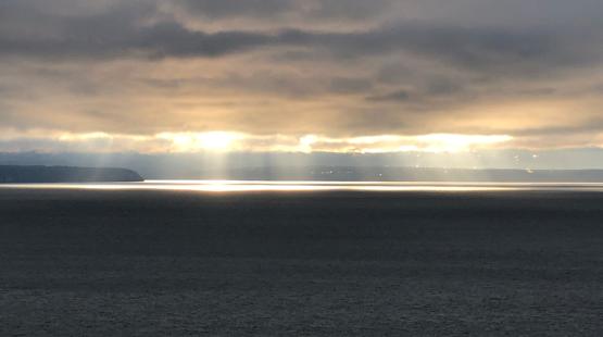 Sunlight breaks through heavy clouds over a wide expanse of water, creating bright reflective bands on the surface. A distant shoreline sits low on the horizon beneath the glowing sky.