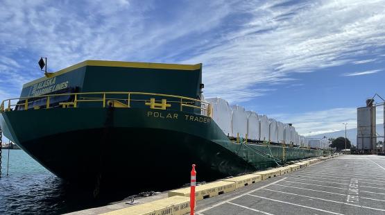 A cargo barge labeled “POLAR TRADER” is docked at a port with white-wrapped freight on deck under a partly cloudy sky.