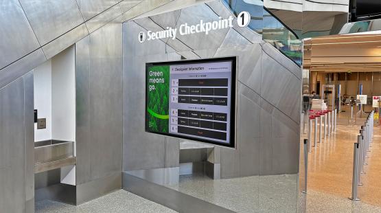 Airport security checkpoint with a digital sign showing lane status and queue stanchions in front.