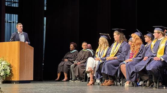 A speaker stands at a podium during a graduation ceremony, while a group of graduates in caps and gowns sit on stage beside several seated faculty or staff members.