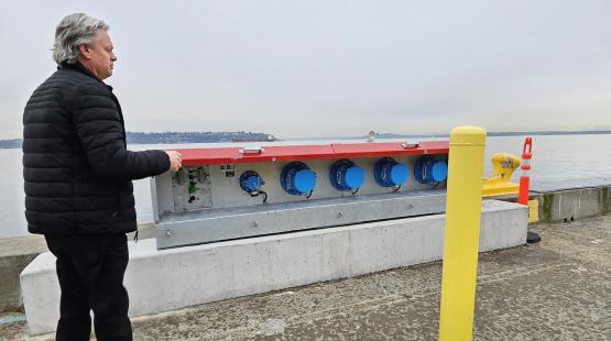 Person standing on a waterfront pier beside a large metal utility box with multiple blue connectors, with water and distant shoreline in the background.