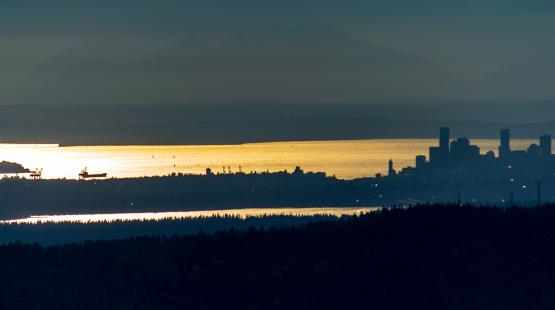 A distant city skyline appears in silhouette above a sunlit bay, with ships and cranes visible along the waterfront.