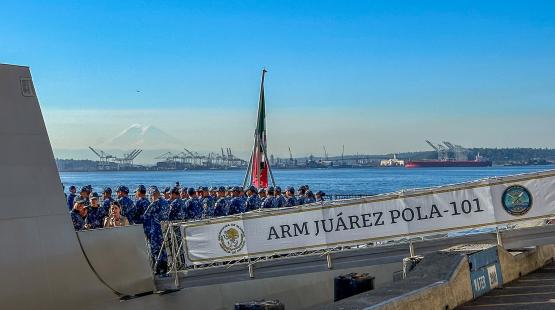 A naval vessel at a waterfront with uniformed crew members gathered on deck near a banner reading “ARM Juárez POLA-101,” with port cranes and a distant mountain in the background.