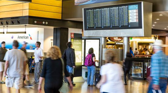 Busy airport concourse with travelers walking and checking a large overhead flight information display.