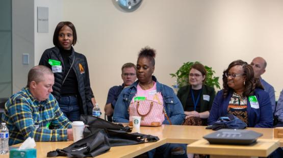 People seated around a meeting table while another person stands and speaks; name tags, drinks, and bags are visible in the room.