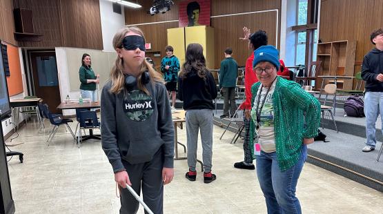 A classroom where a person wearing a blindfold uses a white cane while another person stands nearby, with several others and chairs in the background.