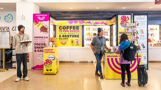 Customers and staff at a brightly colored coffee and gift kiosk in an airport, with signs promoting specialty coffee and products.