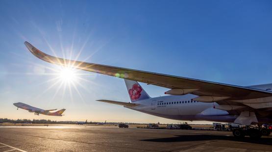A large aircraft sits on the tarmac while another plane takes off in the distance under a bright sun.