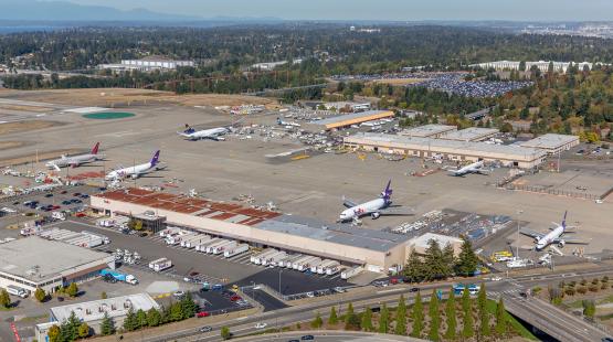A wide aerial view of SEA Airport showing multiple cargo aircraft on the tarmac, airport service vehicles, and surrounding buildings and parking areas.