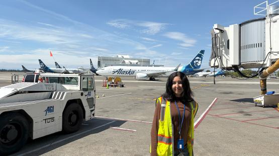 Isabella Newcomer wearing a reflective safety vest stands on an airport tarmac near a jet bridge, with several Alaska‑branded airplanes and ground service vehicles in the background.