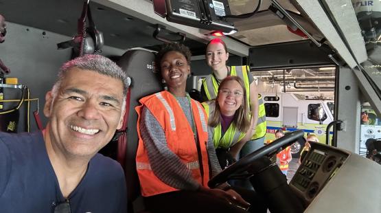 Four people wearing safety vests inside an emergency vehicle cab, with the dashboard and a fire truck visible in the background.