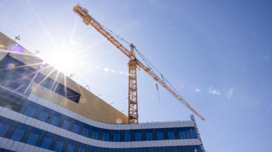 A tall yellow construction crane rises above a modern building with curved blue‑and‑white paneling. Bright sunlight shines behind the crane, creating a strong lens flare across the image. The sky is clear and blue.