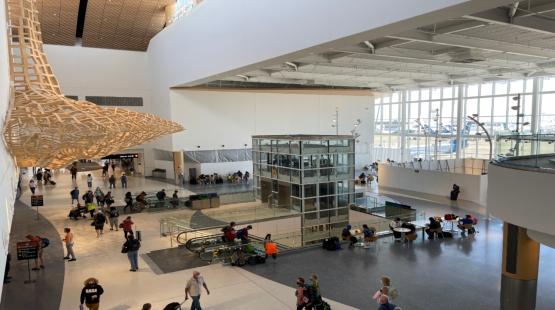 View from the N Concourse showing a large airport hall with travelers, escalators, tall windows facing aircraft, and a suspended wooden art structure.