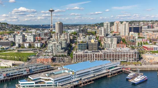 A waterfront cityscape with a long pier building, two docked vessels, and a dense skyline with a tall observation tower in the background.