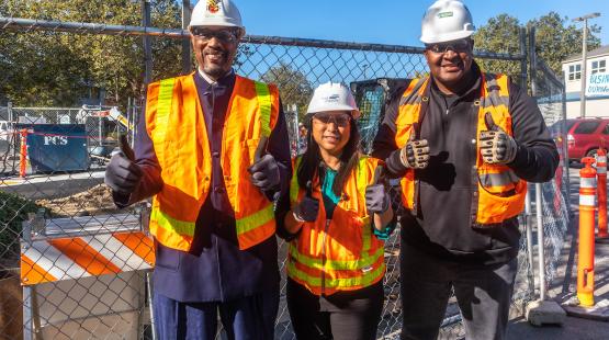 Three construction workers wearing hard hats, safety glasses, gloves, and bright orange reflective vests stand in front of a fenced construction site. All three individuals are giving a thumbs‑up gesture. Behind them are construction materials, vehicles, and trees under a clear blue sky.
