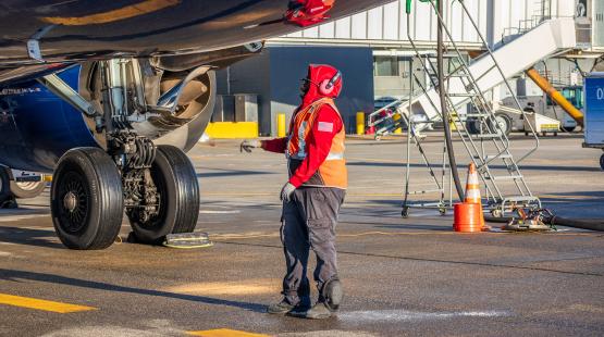An airport ground crew member wearing protective gear and a high‑visibility vest stands near the landing gear of a parked aircraft on the tarmac, inspecting the underside of the plane. Nearby are service equipment, a set of mobile stairs, and traffic cones.