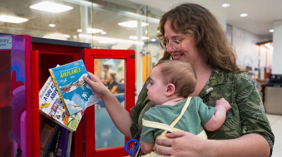 A person holding a young child selects a book from a small red library cabinet filled with children’s books inside an indoor airport area.